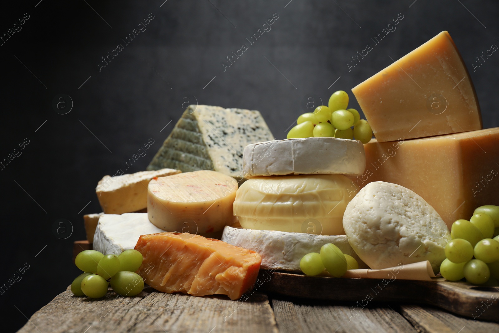 Photo of Different types of cheese and grapes on wooden table against dark background, closeup