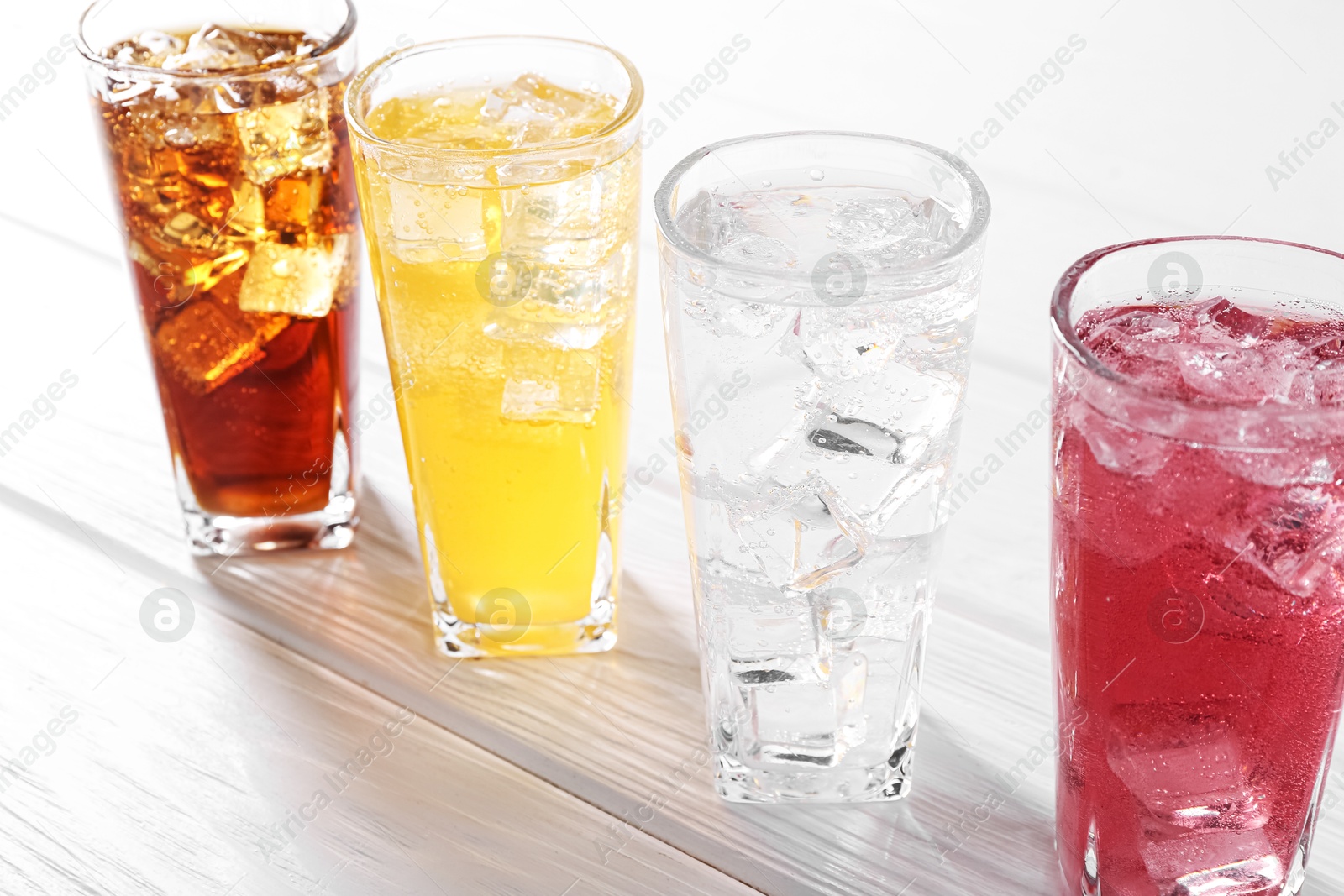 Photo of Soda water of different flavors with ice cubes in glasses on white wooden table, closeup