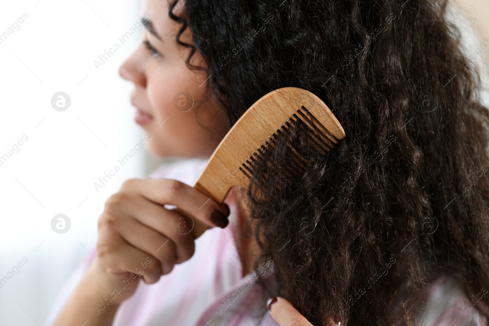 Photo of Young woman brushing her curly hair with comb indoors, closeup. Selective focus