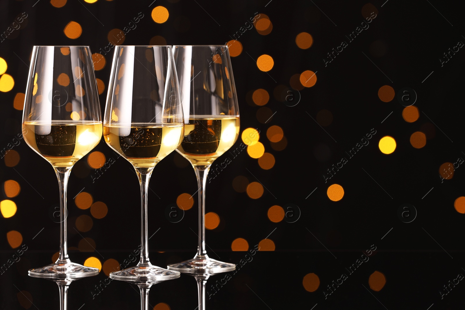 Photo of Tasty white wine in glasses on mirror surface against dark background with blurred lights, space for text. Bokeh effect