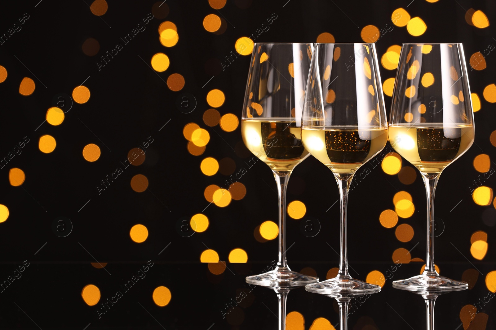 Photo of Tasty white wine in glasses on mirror surface against dark background with blurred lights, space for text. Bokeh effect