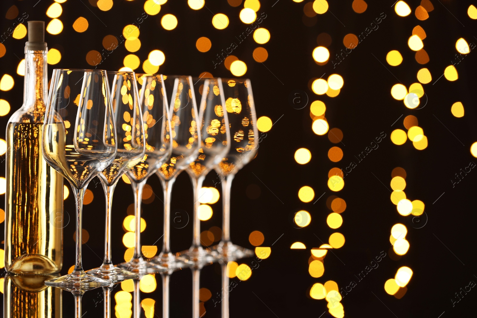 Photo of Tasty white wine in bottle and empty wineglasses on mirror surface against dark background with blurred lights, space for text. Bokeh effect