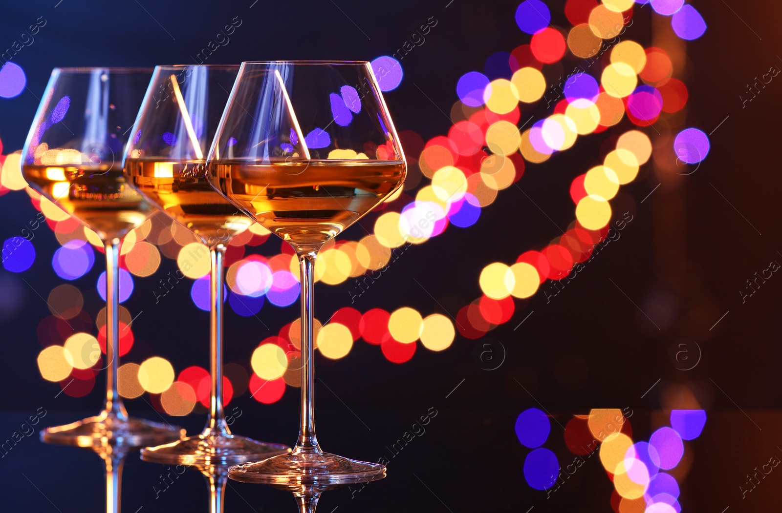 Photo of Tasty white wine in glasses on mirror surface against dark background with blurred lights, space for text. Bokeh effect