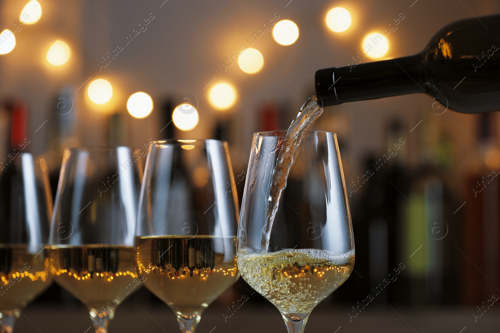 Photo of Pouring white wine into glass against blurred background with festive lights, closeup. Bokeh effect