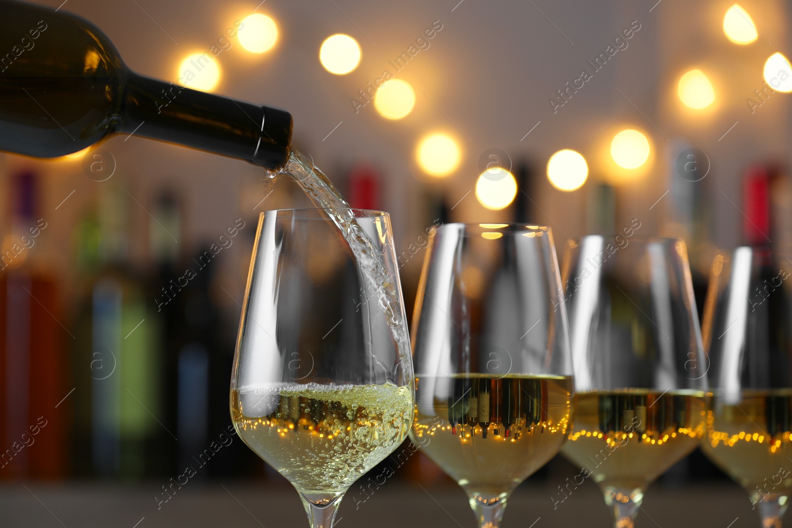 Photo of Pouring white wine into glass against blurred background with festive lights, closeup. Bokeh effect