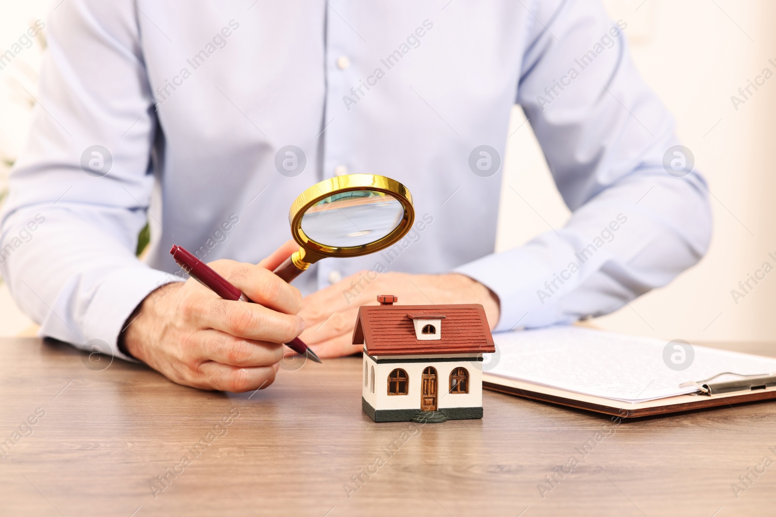 Photo of Real estate agent with pen looking at house figure through magnifying glass at wooden table in office, closeup. Home appraisal