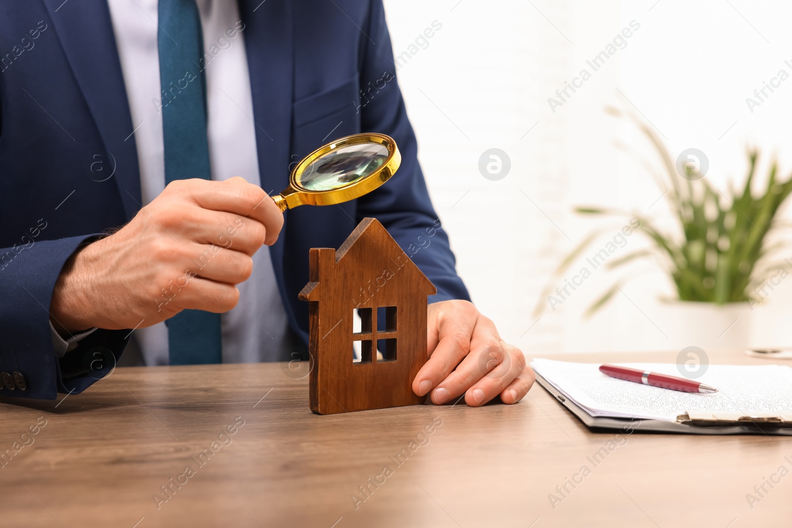 Photo of Real estate agent looking at house figure through magnifying glass at wooden table in office, closeup. Home appraisal