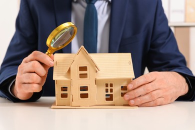 Photo of Real estate agent with house figure and magnifying glass at white table in office, closeup. Home appraisal