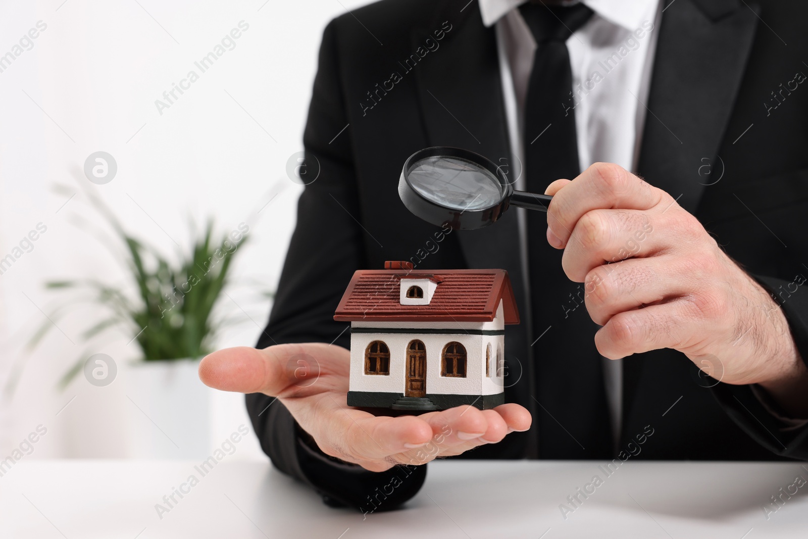 Photo of Real estate agent looking at house figure through magnifying glass at white table in office, closeup. Home appraisal