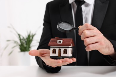 Real estate agent looking at house figure through magnifying glass at white table in office, closeup. Home appraisal