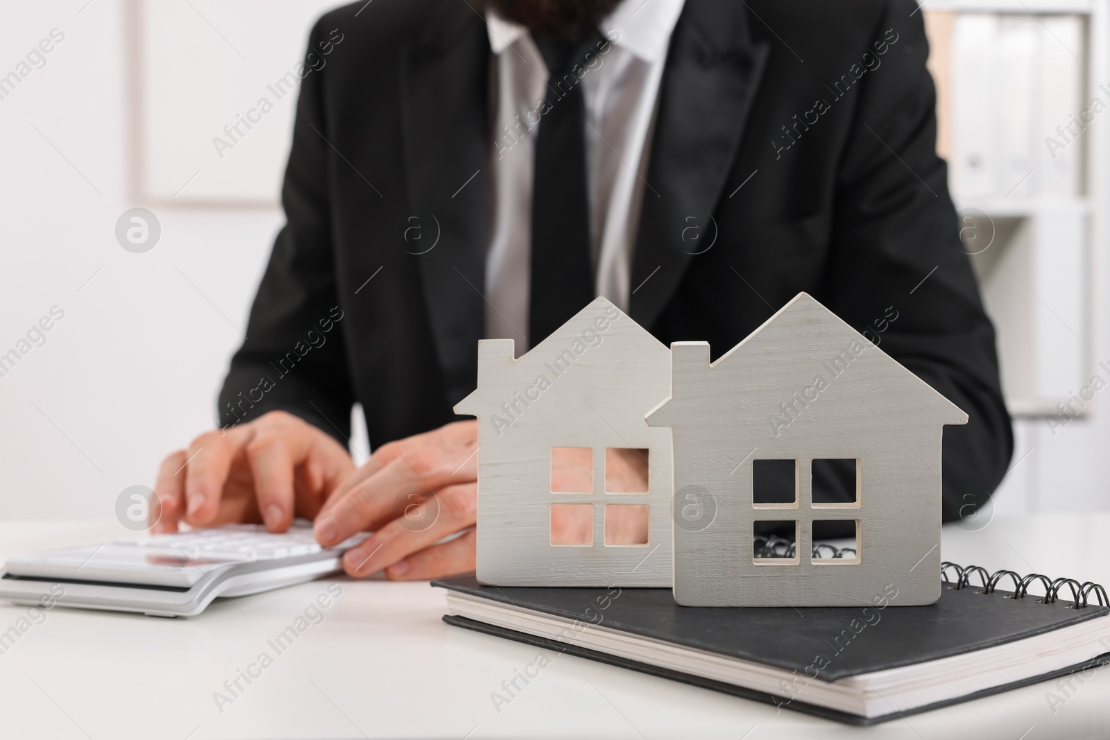 Photo of Real estate agent with calculator at white table in office, focus on house figures and notepad. Home appraisal