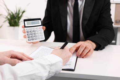 Photo of Real estate agent showing calculator with price for house while client signing document at white table in office, closeup. Home appraisal