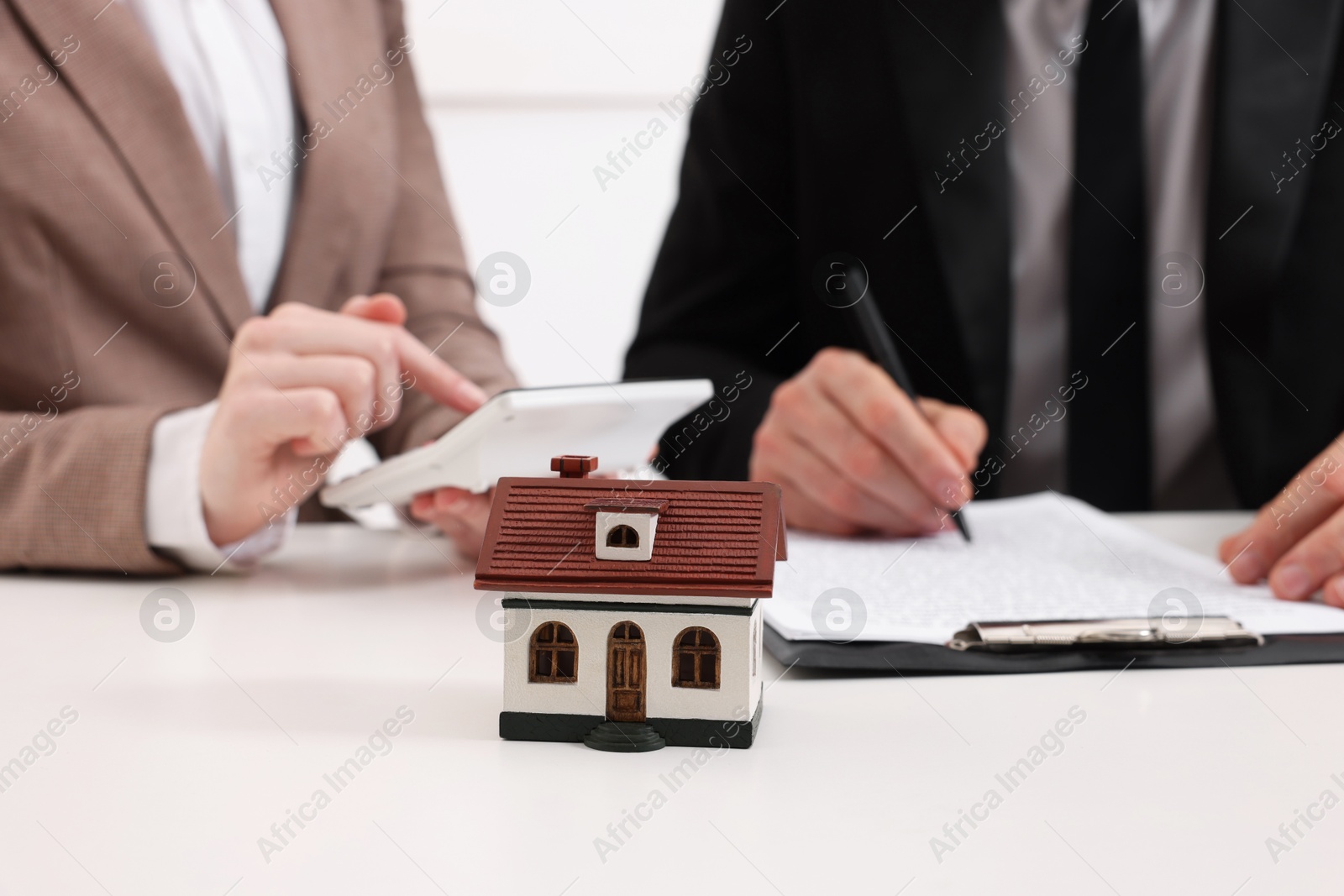 Photo of Real estate agent calculating price for house while client signing document at white table in office, focus on house figure. Home appraisal