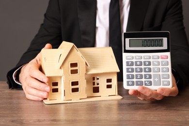 Real estate agent showing calculator with price for house at wooden table against grey background, closeup. Home appraisal