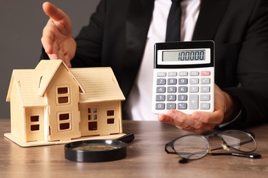 Photo of Real estate agent showing calculator with price for house at wooden table against grey background, closeup. Home appraisal