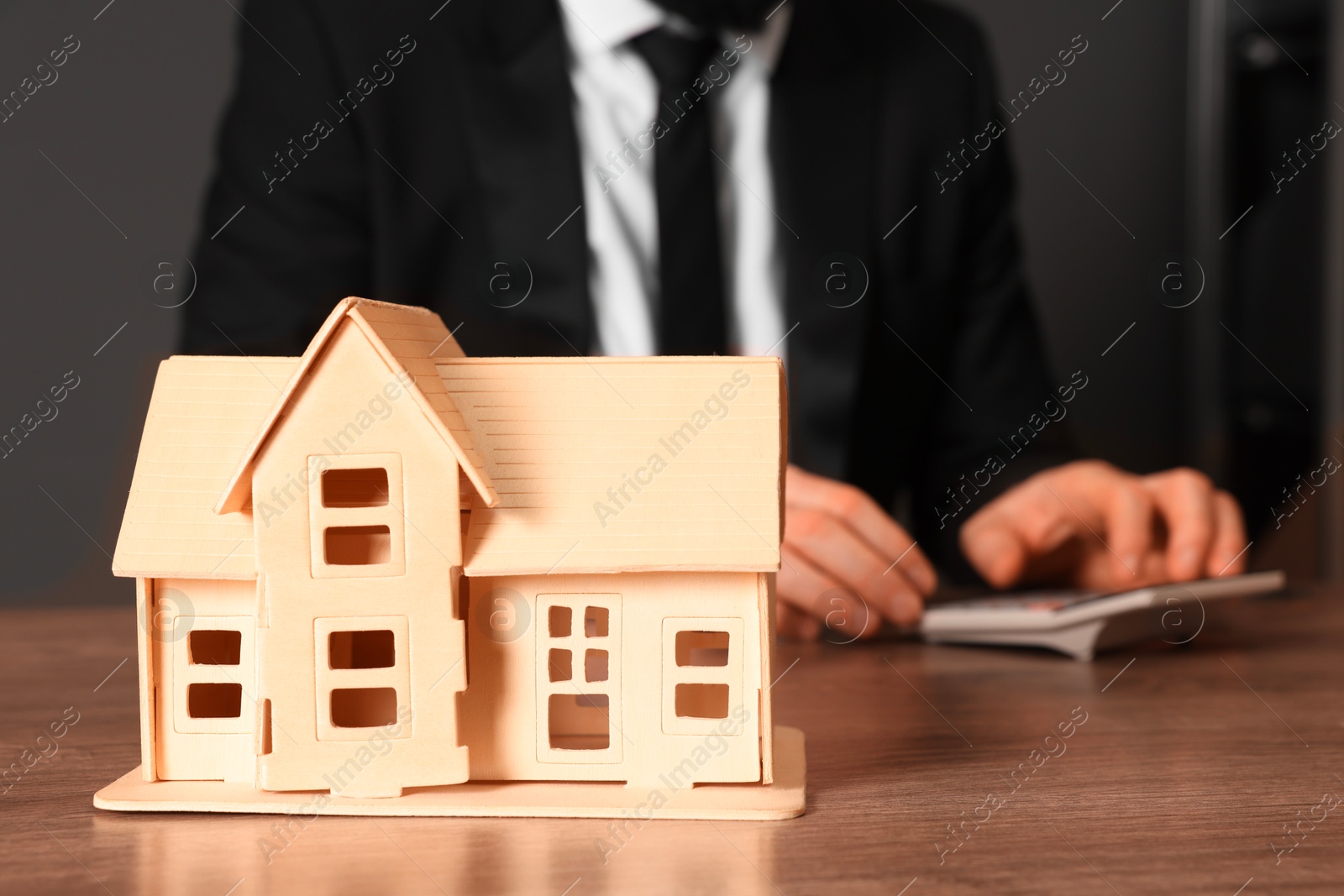 Photo of House figure and real estate agent with calculator at wooden table against grey background, selective focus. Home appraisal