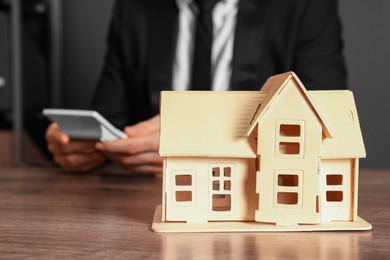 Photo of House figure and real estate agent with calculator at wooden table against grey background, selective focus. Home appraisal