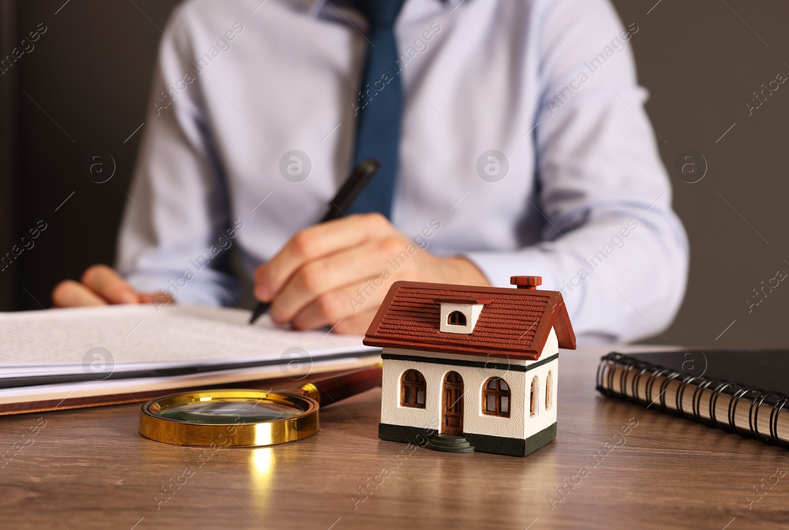 Photo of Real estate agent signing document at wooden table against grey background, focus on house figure and magnifying glass. Home appraisal