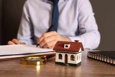 Photo of Real estate agent signing document at wooden table against grey background, focus on house figure and magnifying glass. Home appraisal