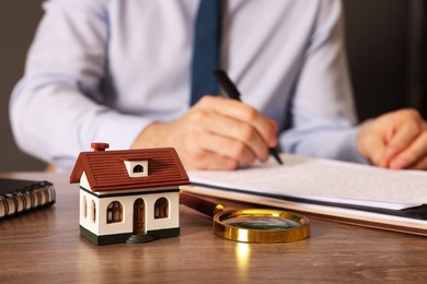 Photo of Real estate agent signing document at wooden table against grey background, focus on house figure and magnifying glass. Home appraisal