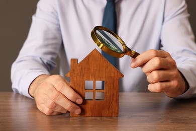 Photo of Real estate agent with house figure and magnifying glass at wooden table against grey background, closeup. Home appraisal