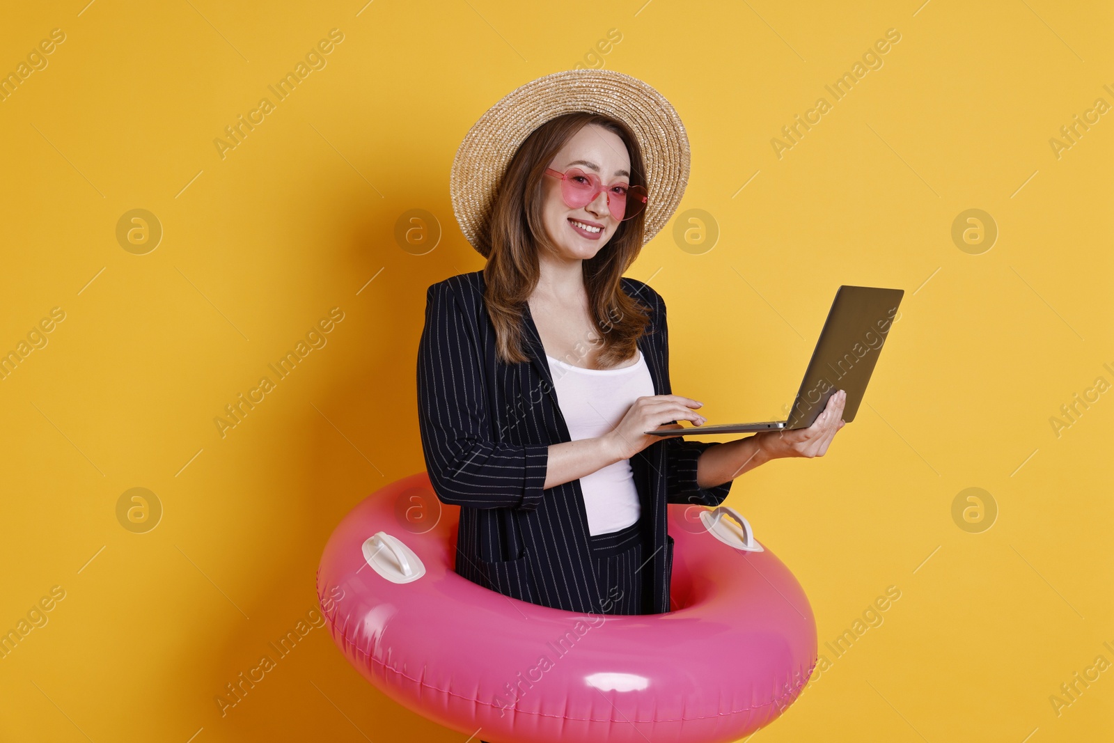 Photo of Businesswoman with inflatable ring, straw hat, sunglasses and laptop on orange background