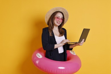 Photo of Businesswoman with inflatable ring, straw hat, sunglasses and laptop on orange background