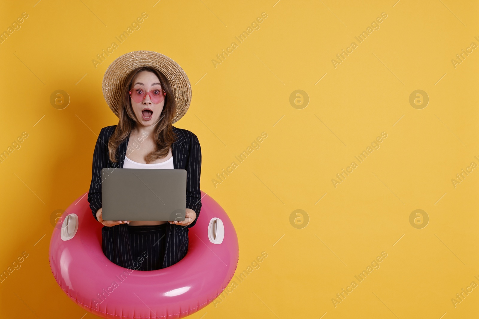 Photo of Surprised businesswoman with inflatable ring, straw hat, sunglasses and laptop on orange background, space for text