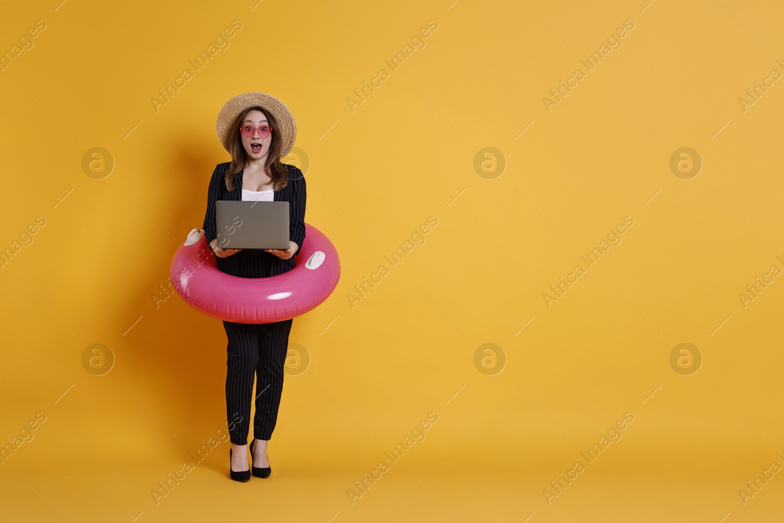 Photo of Surprised businesswoman with inflatable ring, straw hat, sunglasses and laptop on orange background, space for text