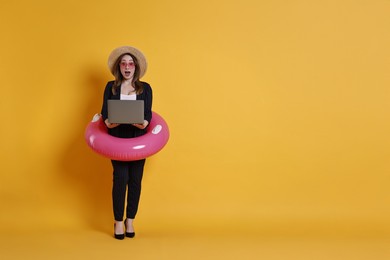 Photo of Surprised businesswoman with inflatable ring, straw hat, sunglasses and laptop on orange background, space for text