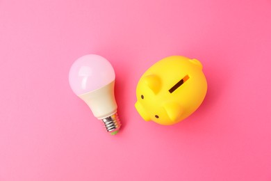 Photo of Piggy bank and light bulb on pink background, flat lay. Energy saving concept