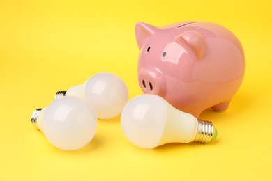 Photo of Piggy bank and light bulbs on yellow background, closeup. Energy saving concept