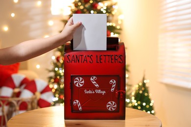 Photo of Little child putting letter to Santa Claus into red mail box indoors, closeup. Christmas tradition