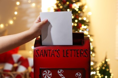 Photo of Little child putting letter to Santa Claus into red mail box indoors, closeup. Christmas tradition