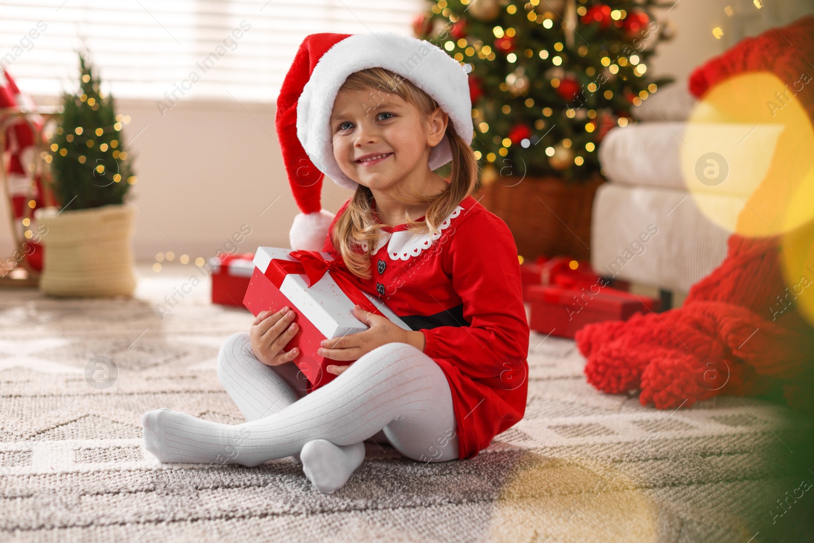 Photo of Little girl in Santa hat with gift box sitting on floor at home