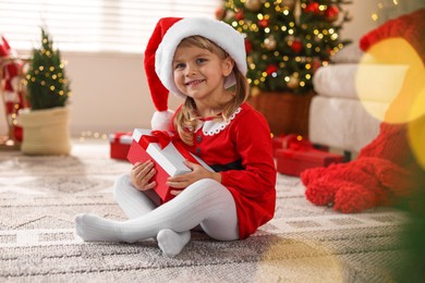 Photo of Little girl in Santa hat with gift box sitting on floor at home