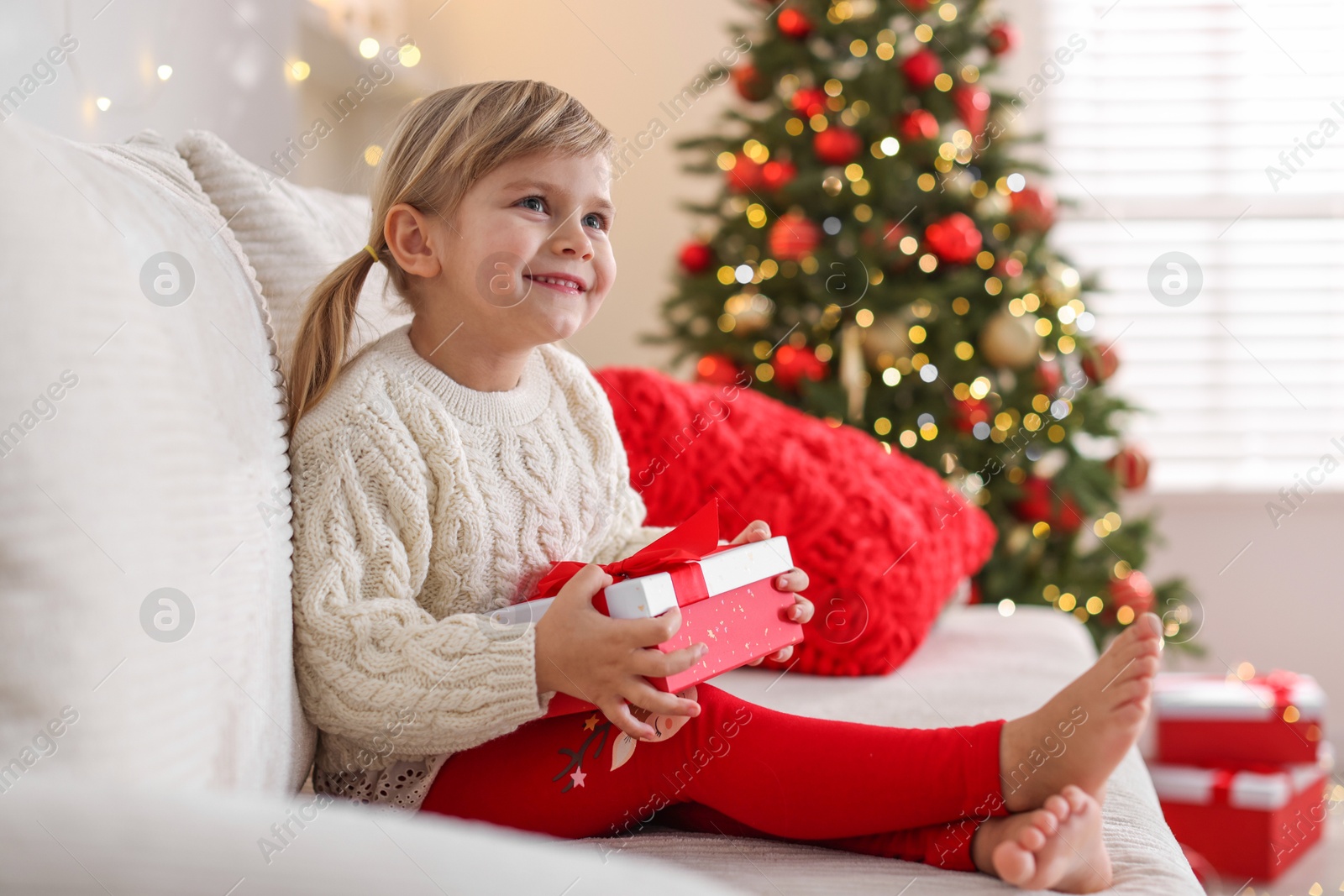 Photo of Little girl with Christmas gift box sitting on sofa at home