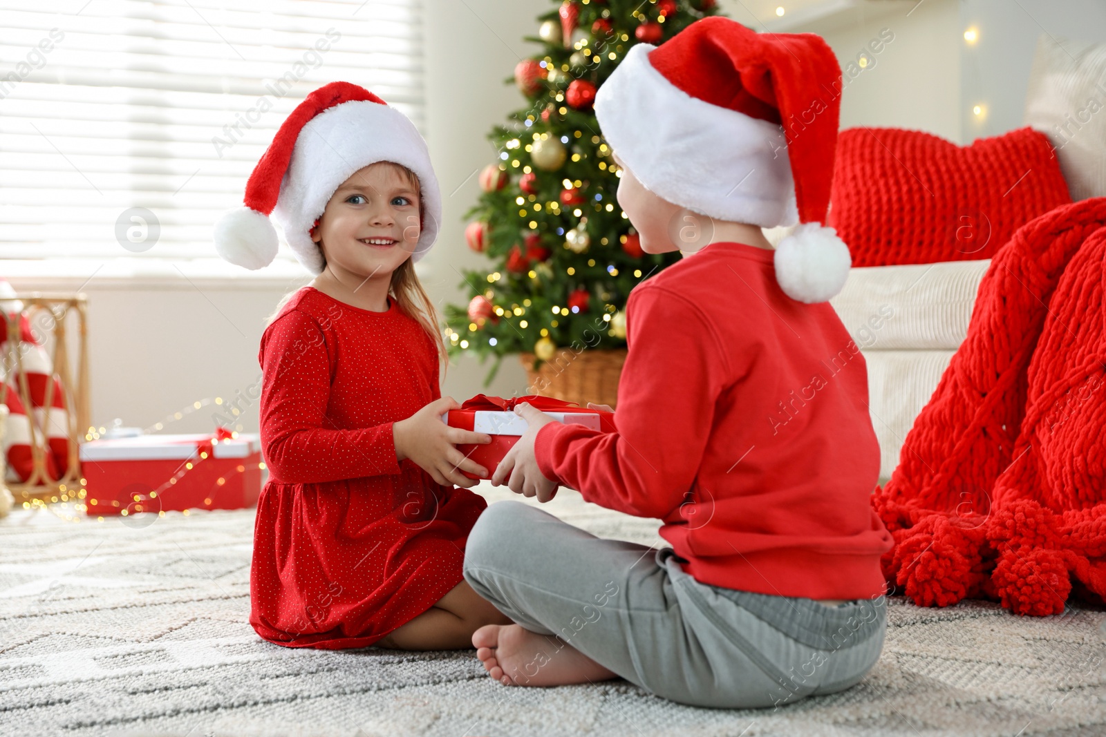 Photo of Happy little girl in Santa hat presenting her brother with Christmas gift at home