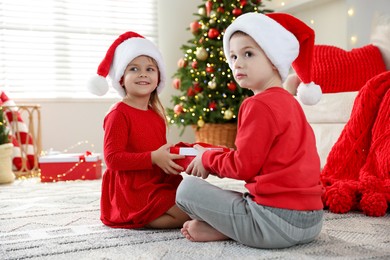 Photo of Happy little girl in Santa hat presenting her brother with Christmas gift at home