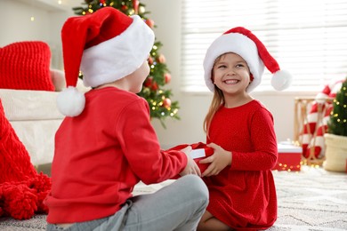 Photo of Happy little girl in Santa hat presenting her brother with Christmas gift at home