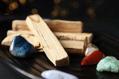 Photo of Palo santo sticks and gemstones on black table against blurred lights, closeup