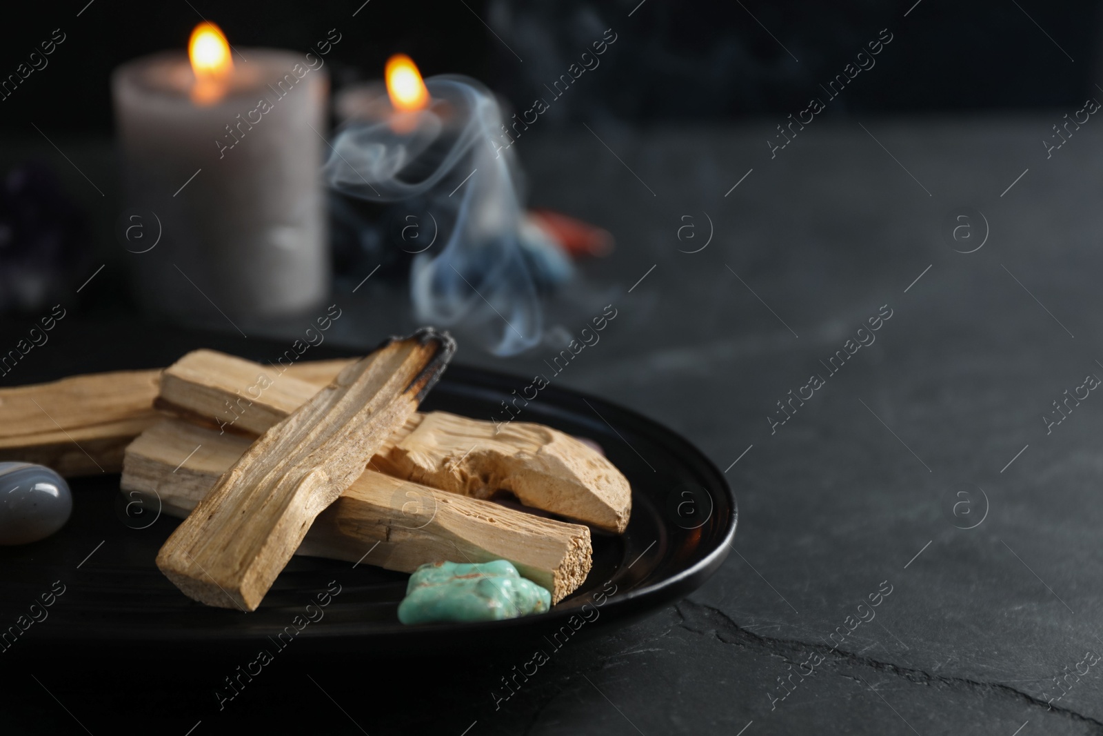 Photo of Smoldering palo santo stick, gemstones and burning candles on black table, closeup. Space for text