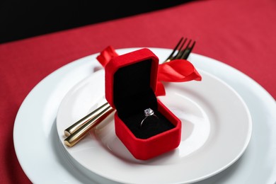 Photo of Beautiful engagement ring and setting for romantic dinner on red table, closeup