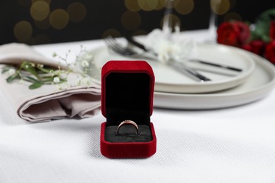 Photo of Engagement ring in box, flowers and tableware on white tablecloth, closeup. Romantic dinner