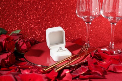 Photo of Engagement ring in box, tableware, rose flowers and petals on red glitter background, closeup. Table setting for romantic dinner