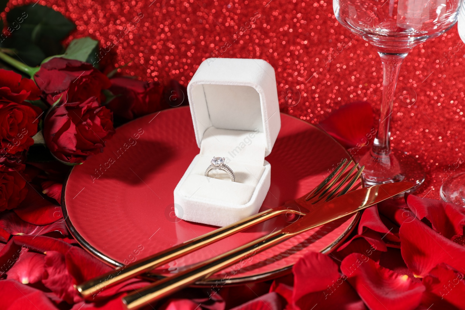 Photo of Engagement ring in box, tableware, rose flowers and petals on red glitter background, closeup. Table setting for romantic dinner