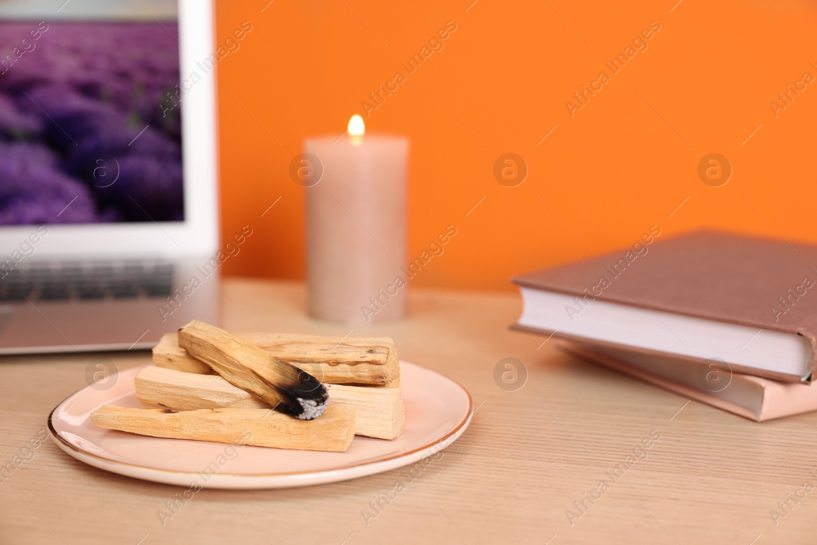 Photo of Palo santo sticks, laptop, books and burning candle on wooden table against orange wall