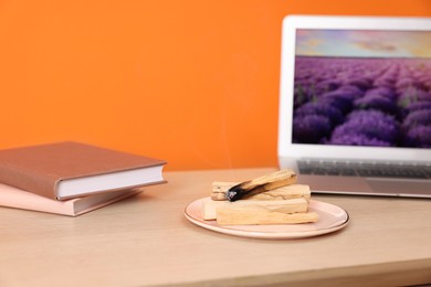 Photo of Palo santo sticks, laptop and books on wooden table against orange wall, space for text