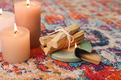 Photo of Palo santo sticks and burning candles on rug indoors, closeup