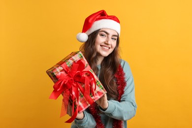 Photo of Happy young woman with tinsel, Santa hat and gift box on orange background